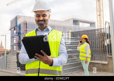 Engineer uses a tablet on a construction site while a colleague talks on the phone. Both wear safety gear and reflect the modern construction working Stock Photo
