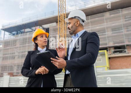 Two engineers in hard hats discuss a construction project on site. They hold a tablet and documents, collaborating on building development in an urban Stock Photo