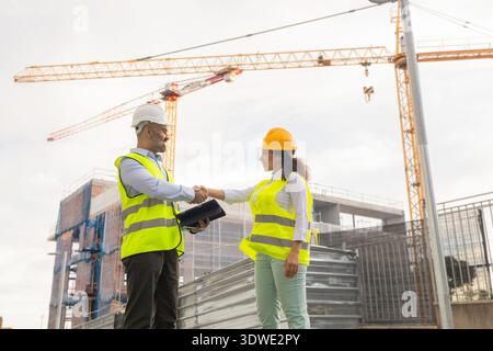 Two engineers in safety gear shake hands on a construction site, symbolizing successful collaboration. Behind them, cranes and a building under constr Stock Photo