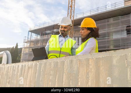 Engineers in safety vests and helmets review plans on a tablet at a building site, collaborating to ensure project success and structural integrity. Stock Photo