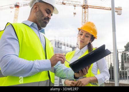 Engineers in safety gear review blueprints on a tablet at a construction site. Cranes and a building structure are seen in the background, emphasizing Stock Photo