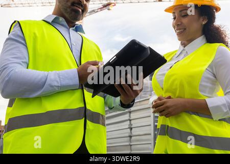 Engineers review project plans on a tablet at a building site. They are wearing high-visibility vests and hard hats to ensure safety while discussing Stock Photo