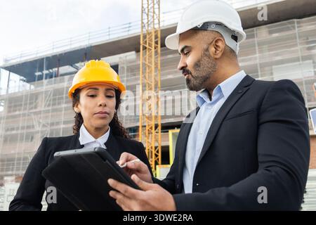 Two engineers wearing helmets discuss project plans on a construction site. They use a digital tablet to review blueprints, reflecting modern construc Stock Photo