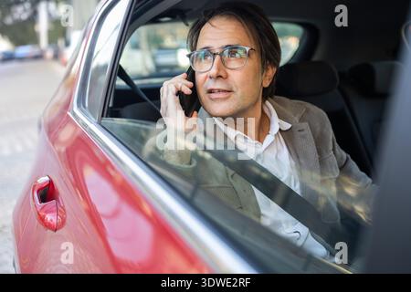 A man in a car engaged in a conversation on his smartphone. He gazes forward, possibly planning his journey or discussing travel details during a road Stock Photo