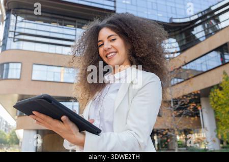 A young Afro Venezuelan woman with curly hair, wearing a professional white suit, smiles confidently while using a tablet outside a modern office buil Stock Photo