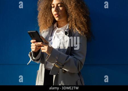 Afro Venezuelan woman with curly hair standing against a blue wall, focused on her phone. She wears a gray coat and a white shirt, embodying modern li Stock Photo