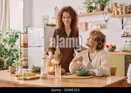A redhead woman serves breakfast to her blonde partner in their stylish home. Stock Photo