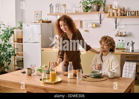 Redhead woman serves breakfast to her blonde partner in their stylish kitchen setting. Stock Photo