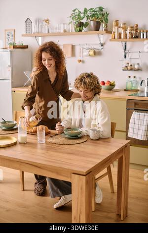 In a cozy modern kitchen, a redhead woman serves breakfast to her blonde partner at the table. Stock Photo