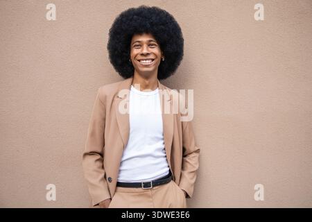 A person stands confidently against a neutral background, wearing a stylish brown blazer and a casual white top, exuding warmth and positivity with a Stock Photo