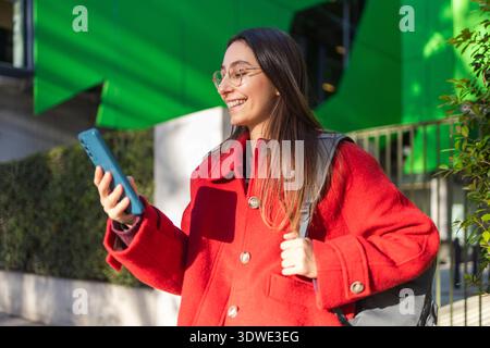 A smiling woman in a bright red coat stands outside during winter, holding her smartphone and enjoying the crisp air. She appears content and stylish Stock Photo