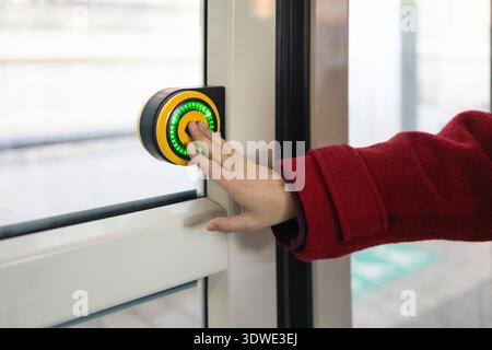A woman in a red coat presses a green lit button on a bus door in winter. The scene captures a moment of interaction with public transport, showcasing Stock Photo