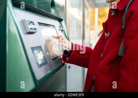 A woman wearing a vibrant red coat utilizes a touchscreen interface outdoors during winter. The scene captures the blend of technology with seasonal f Stock Photo
