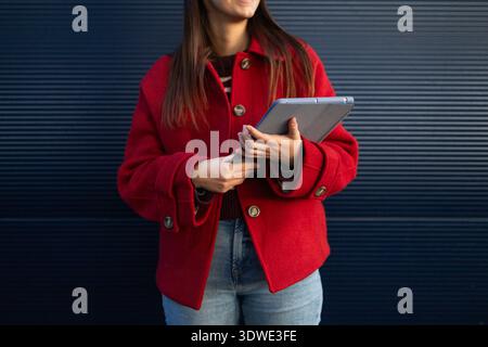 A woman in a stylish red coat stands against a dark background, holding a tablet in her hands. The cozy garment highlights the winter season, blending Stock Photo