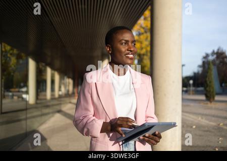African businesswoman in a pink blazer smiles confidently while using a digital tablet outside a modern office building, portraying successful leaders Stock Photo