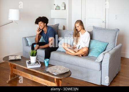 A multiethnic couple enjoys leisure time in a cozy living room. The man talks on the phone while the woman uses a tablet, both sitting on a comfortabl Stock Photo