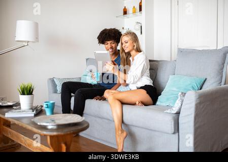 A joyful multiethnic couple enjoys leisure time together, using a tablet while seated comfortably on a stylish gray couch in a modern living room sett Stock Photo
