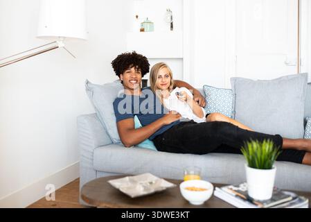 A multiethnic couple is relaxing comfortably on a cozy sofa in their modern living room, enjoying some leisure time together while watching TV at home Stock Photo