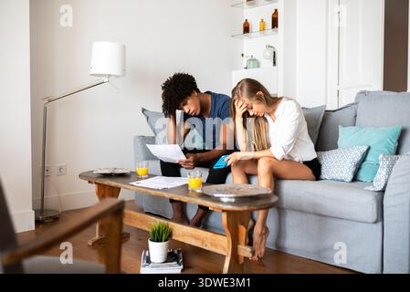 A multiethnic couple sits on a couch in their home, visibly stressed while reviewing financial documents. The atmosphere suggests concern and the chal Stock Photo