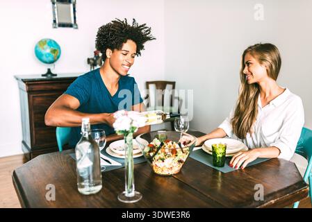 A multiethnic couple shares a meal at a cozy home dining table, creating a warm and welcoming atmosphere. The young man pours wine while his partner s Stock Photo