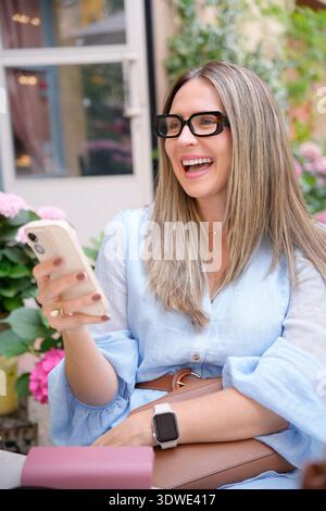 Smiling mature woman with eyeglasses using a smartphone in a cafe. She wears a light blue blouse, holding her mobile phone and a bag, enjoying her tim Stock Photo