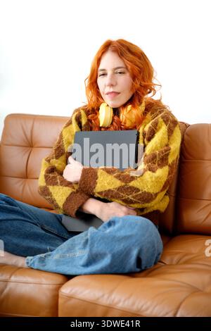 A woman with red hair sits comfortably on a brown leather sofa, wearing a cozy yellow sweater and blue jeans, holding a digital tablet and sporting br Stock Photo