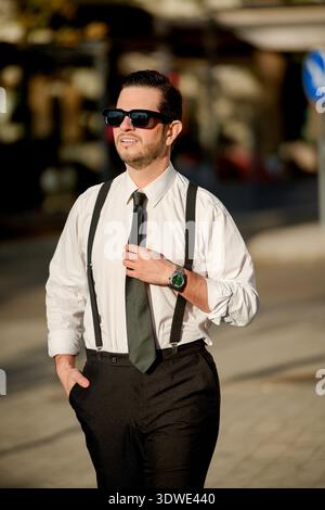 Confident businessman in formal attire, wearing sunglasses, walks outdoors in sunny weather. His attire includes a tie, suspenders, and a stylish watc Stock Photo