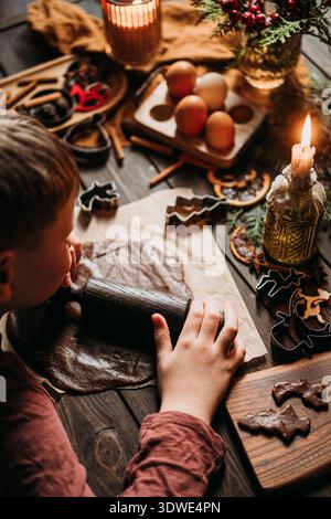 A child is joyfully rolling out gingerbread dough on a wooden table, surrounded by various baking ingredients and cookie cutters, creating festive hol Stock Photo