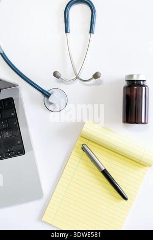 Top view of a medical office workspace featuring a laptop, stethoscope, pharmaceutical bottle, and a pen on a notepad, suggesting a professional healt Stock Photo