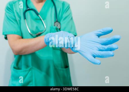 Cropped unrecognizable image of a female healthcare professional wearing a green medical uniform and putting on protective blue gloves. Focus on the a Stock Photo
