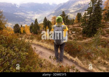 A hiker walks along a scenic trail in the Alps, surrounded by vibrant autumn foliage and towering mountains, capturing the beauty of the season beneat Stock Photo