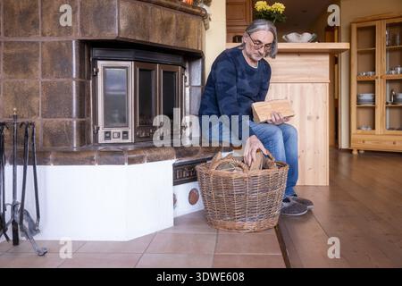 A man selects firewood from a wicker basket to place in a tiled fireplace. The warm, wooden interior and cozy slippers enhance the homely, rustic feel Stock Photo