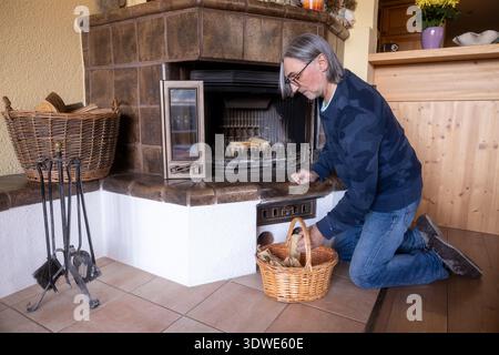 An adult man is seen preparing a home fireplace for lighting. He is kneeling down, organizing wooden logs to ensure a warm and inviting atmosphere in Stock Photo