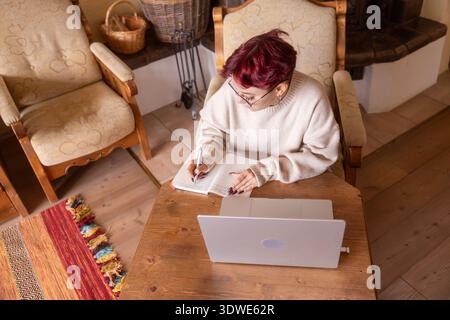 A young woman with glasses sits comfortably in a cozy living room, writing notes in a notebook while working on her laptop. The setting suggests a pro Stock Photo