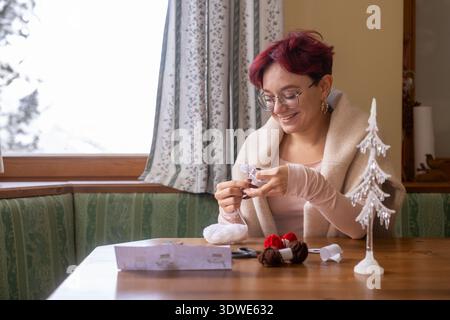 A young woman with short red hair and glasses enjoys crafting at her cozy home. Sitting by a window, she focuses on her project, surrounded by supplie Stock Photo