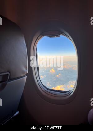 A serene view from an airplane window showcasing a beautiful sunset over fluffy clouds. The sky is painted in soft hues of orange and blue, providing Stock Photo