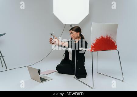 A woman sits on a studio floor, smiling as she takes a selfie with her smartphone. Bright lighting and a laptop behind her create a professional envir Stock Photo