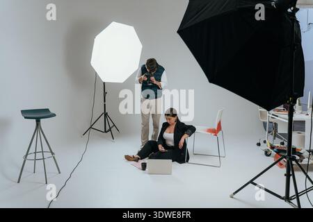Photographer adjusting camera settings behind the scenes in a studio. Model seated with a laptop on the floor, creating a dynamic work environment atm Stock Photo
