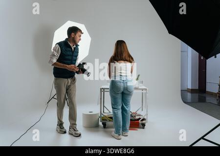 Studio setup showing a photographer holding a camera, engaging with a model standing by a table. The scene captures behind-the-scenes dynamics of a pr Stock Photo