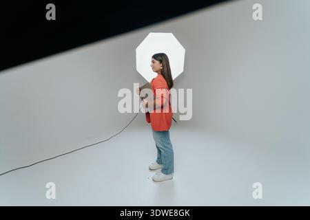 Woman stands confidently in a studio setting, holding a folder. A large photography light is illuminated behind her, showcasing a professional environ Stock Photo