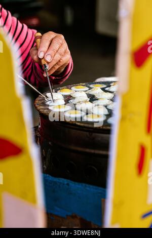 Hand of a street vendor skillfully cooking quail eggs on a hot plate at a bustling market in Taiwan, showcasing the vibrant and diverse food culture o Stock Photo