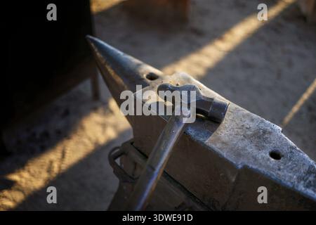A detailed view of a blacksmith’s hammer resting on an anvil in a medieval sword forge. The ambient light highlights the textures of the metal, evokin Stock Photo