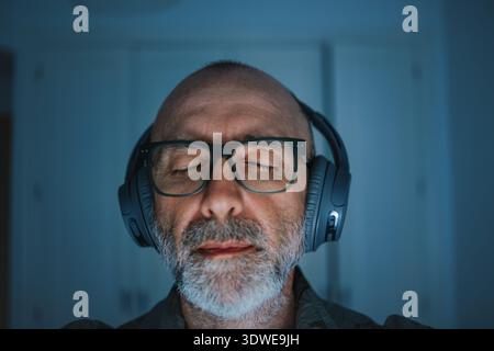 A middle-aged man with glasses is deeply immersed in music, wearing headphones, and has his eyes closed. The scene conveys relaxation and enjoyment in Stock Photo