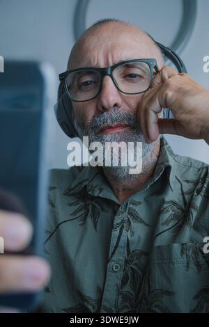 A middle-aged man wearing headphones is deeply immersed in music. Seated comfortably, he focuses on his smartphone, enjoying the auditory experience i Stock Photo