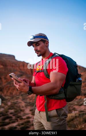 A man hikes through Spain's Red Canyon, using his smartphone to navigate the stunning landscape. He is equipped with a headlamp and a backpack, emphas Stock Photo