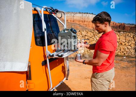 A young man in a red shirt prepares for a canyon adventure in Spain, using a portable container on a vibrant orange van against a backdrop of striking Stock Photo