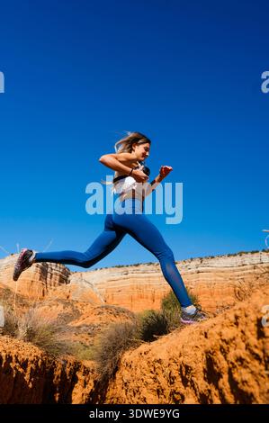 A woman captures the spirit of adventure as she leaps across a gap in the stunning Red Canyon landscape, her camera in hand, perfectly blending explor Stock Photo