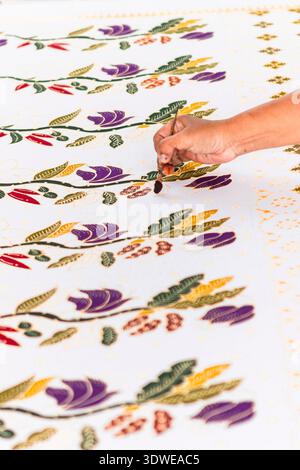 A close-up of a skilled artisan's hand applying traditional Indonesian batik hand-dyeing techniques on fabric. The intricate patterns feature vibrant Stock Photo