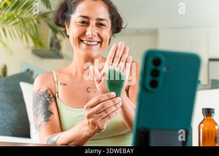 A woman demonstrates a self-care skincare routine using a Gua Sha tool at home. She smiles while recording a video tutorial, emphasizing relaxation an Stock Photo