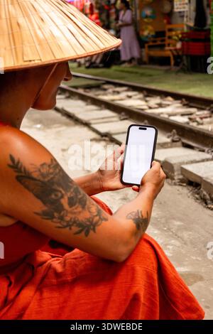 Person wearing a traditional Vietnamese conical hat, known as non la, holding a modern smartphone with a blank screen, sitting by train tracks in Hano Stock Photo